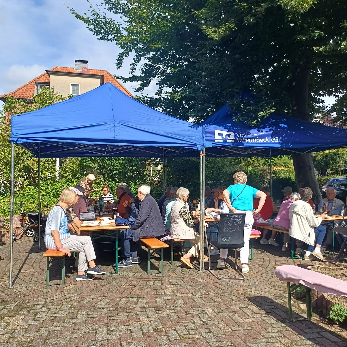 Die Pavillons, die nun Schutz vor der heißen Sonne bieten. Darunter stehen Tische und Bänke und alle Plätze sind belegt mit Gästen, die das Sommerfest wirklich genießen können.