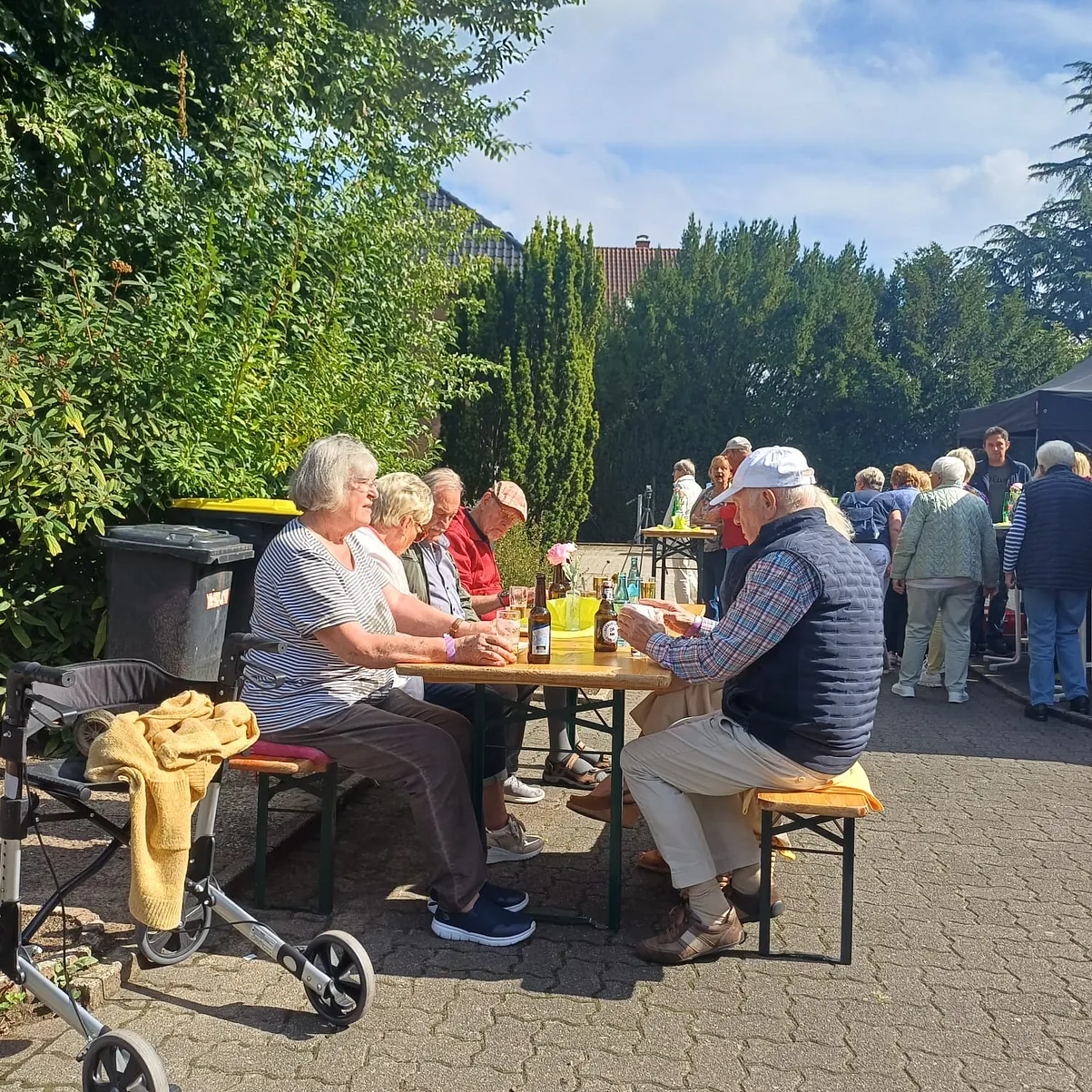 Der Blick ist im freien, in der Sonne auf einige Sommerfestgäste, auf Bänke vor einen Tisch. Weiter im Hintergrund kann man noch die Schlange sehen, wo sich die Gäste für das Essen anstellen.