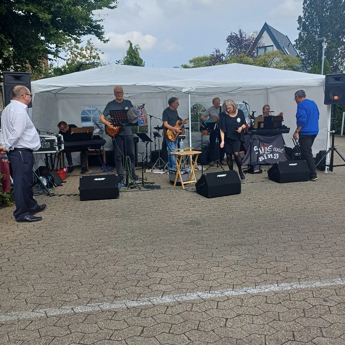 Der Blick auf die Band. Man sieht die Mitglieder an ihren Musikinstrumenten und die Sängerin, sowie auf der Seite ein Gast, der sich das ansieht und vor allen anhört.